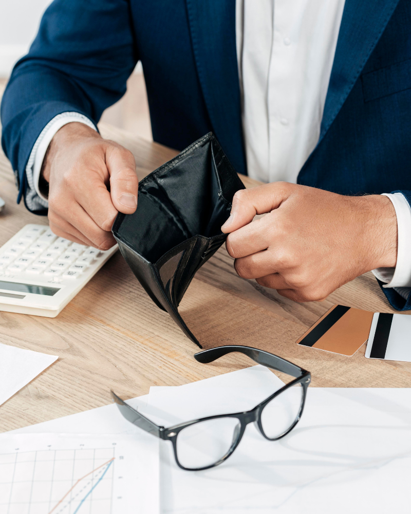 manos de hombre vestido formal abriendo billetera vacía sobre un escritorio con calculadora documentos y tarjetas